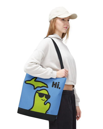 Woman holding a 16"x16" Michigan tote bag with a green Michigan-shaped character wearing sunglasses and smiling and saying "Hi" on a blue background.