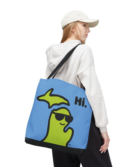 Woman holding a 18"x18" Michigan tote bag with a green Michigan-shaped character wearing sunglasses and smiling and saying "Hi" on a blue background.
